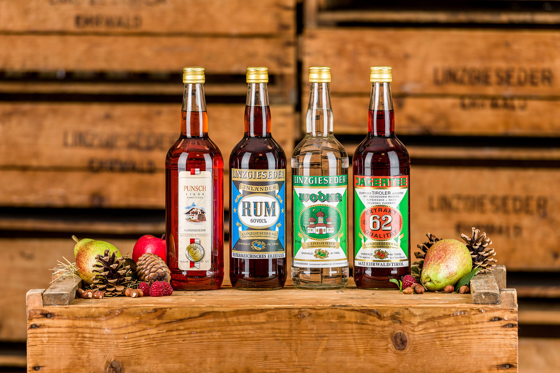 Vier Linzgieseder Spirituosenfalschen auf einer Holzkiste stehend, von links nach rechts: Linzgieseder Punschlikör, Inländer Rum 60%, Jagertee 22,5% und Inländer Rum 62%. Links und rechts daneben liegen Äpfel, Birnen, Nüsse und Zapfen. Der Hintergrund zei