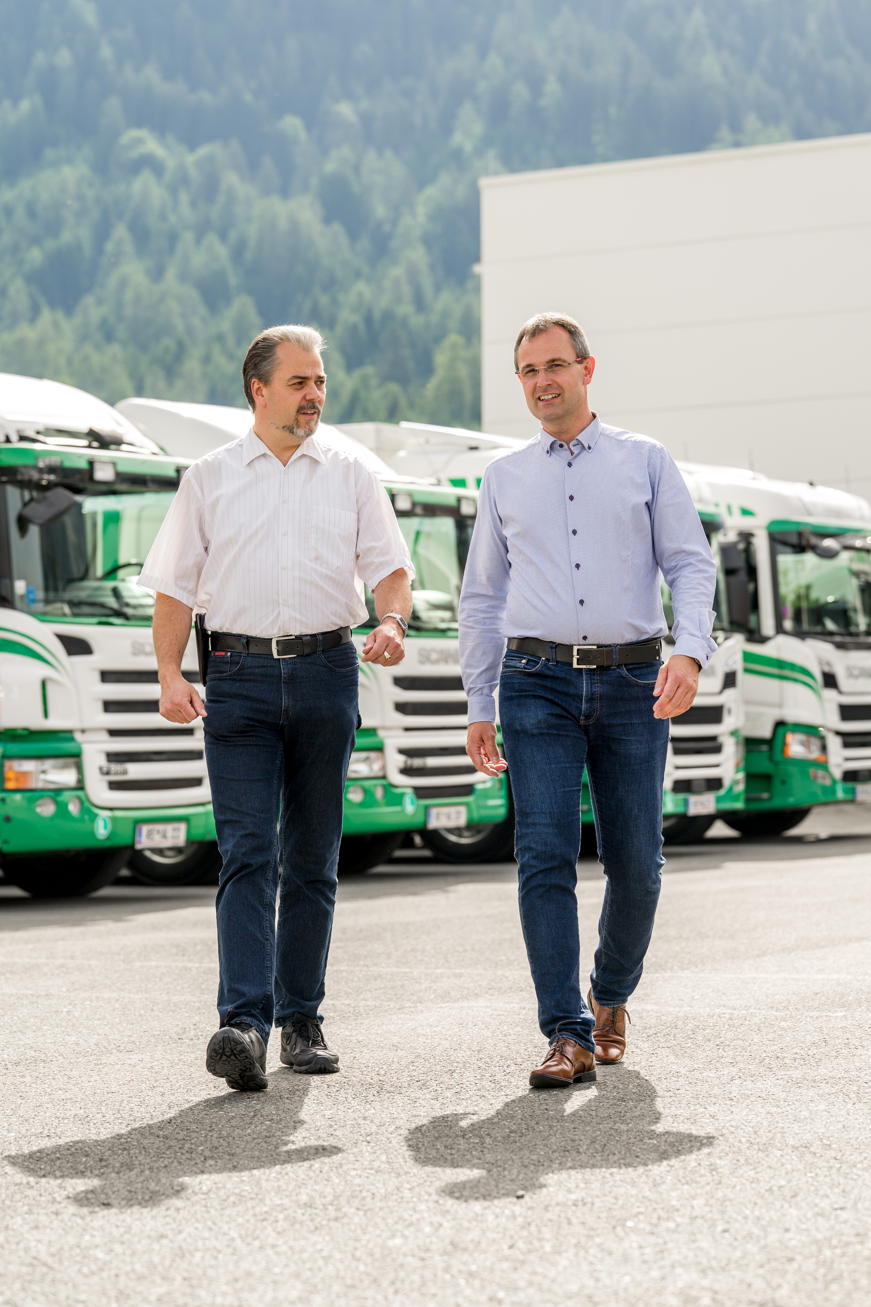 Arthur und Alfred Linzgieseder gehen über das Betriebsgelände. Im Hintergrund parken Lkw der Firma, die ordentlich auf dem Gelände abgestellt sind. Die beiden Männer sind in Bewegung, während die Lkw ruhig im Hintergrund stehen.