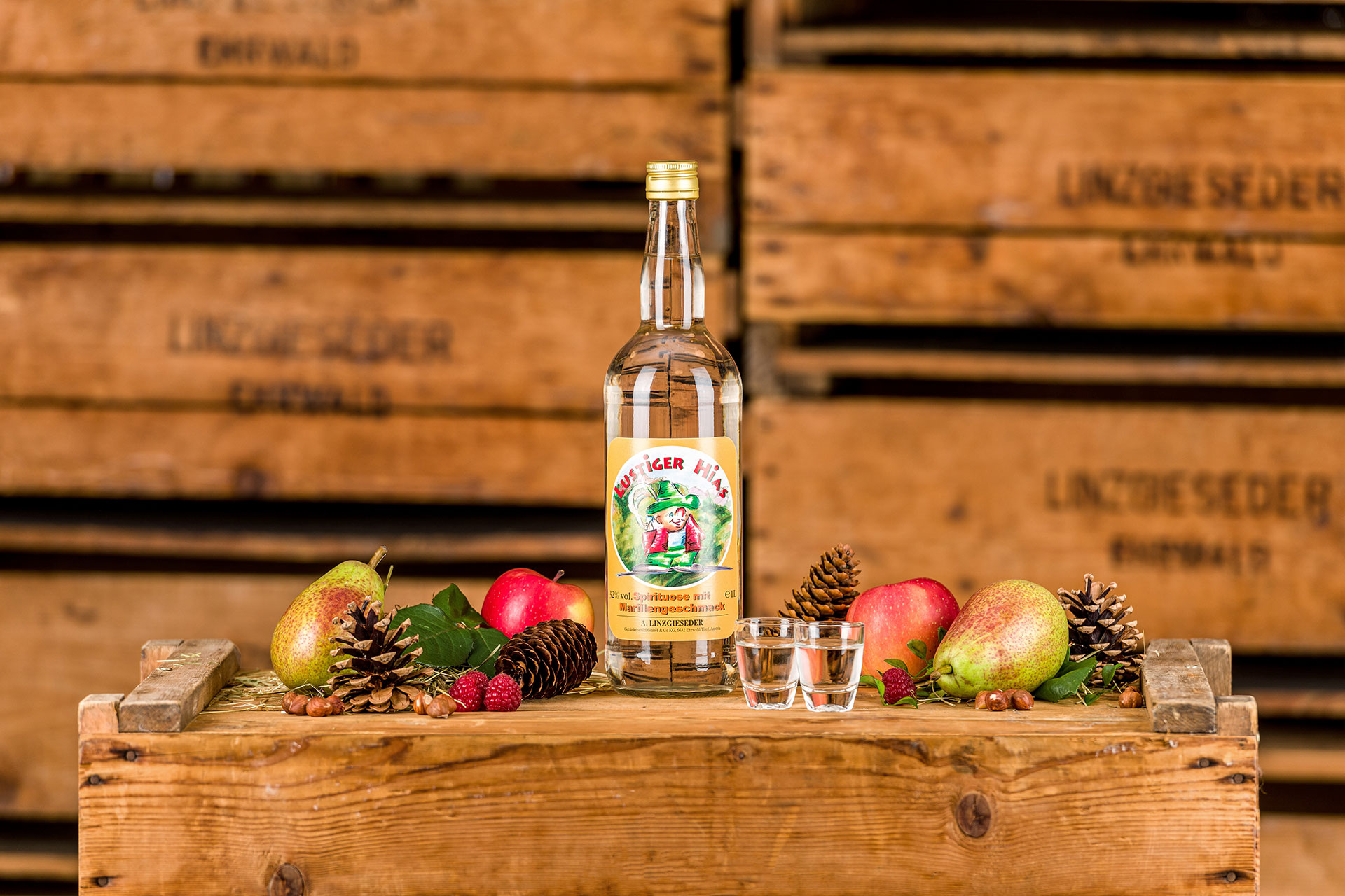 Spirituosenflasche ‚Lustiger Hias Marille‘ mit zwei gefüllten Schnapsgläsern mittig auf einer Holzkiste stehend. Links und rechts davon liegen Birnen, Äpfel, Zapfen und Nüsse. Der Hintergrund zeigt gestapelte Holzkisten mit Linzgieseder-Aufschrift.