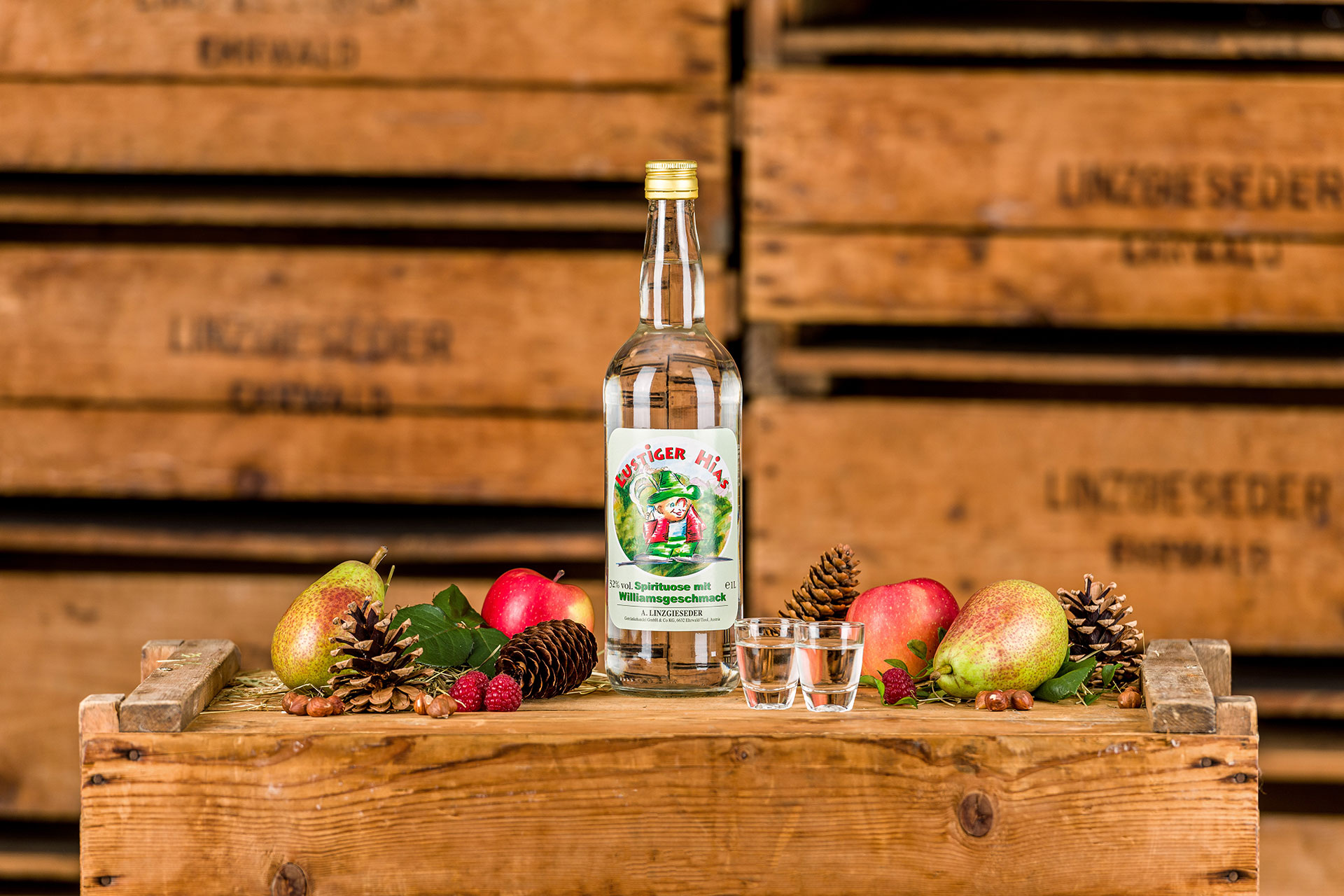 Spirituosenflasche ‚Lustiger Hias Williamsbirne‘ mit zwei gefüllten Schnapsgläsern mittig auf einer Holzkiste stehend. Links und rechts davon liegen Birnen, Äpfel, Zapfen und Nüsse. Der Hintergrund zeigt gestapelte Holzkisten mit Linzgieseder-Aufschrift.