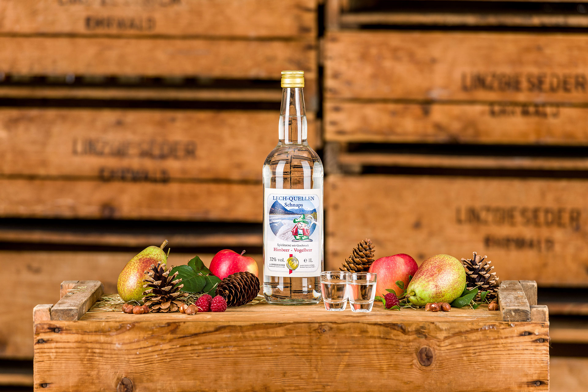 Spirituosenflasche ‚Lustiger Hias Lechquelle‘ mit zwei gefüllten Schnapsgläsern mittig auf einer Holzkiste stehend. Links und rechts davon liegen Birnen, Äpfel, Zapfen und Nüsse. Der Hintergrund zeigt gestapelte Holzkisten mit Linzgieseder-Aufschrift.