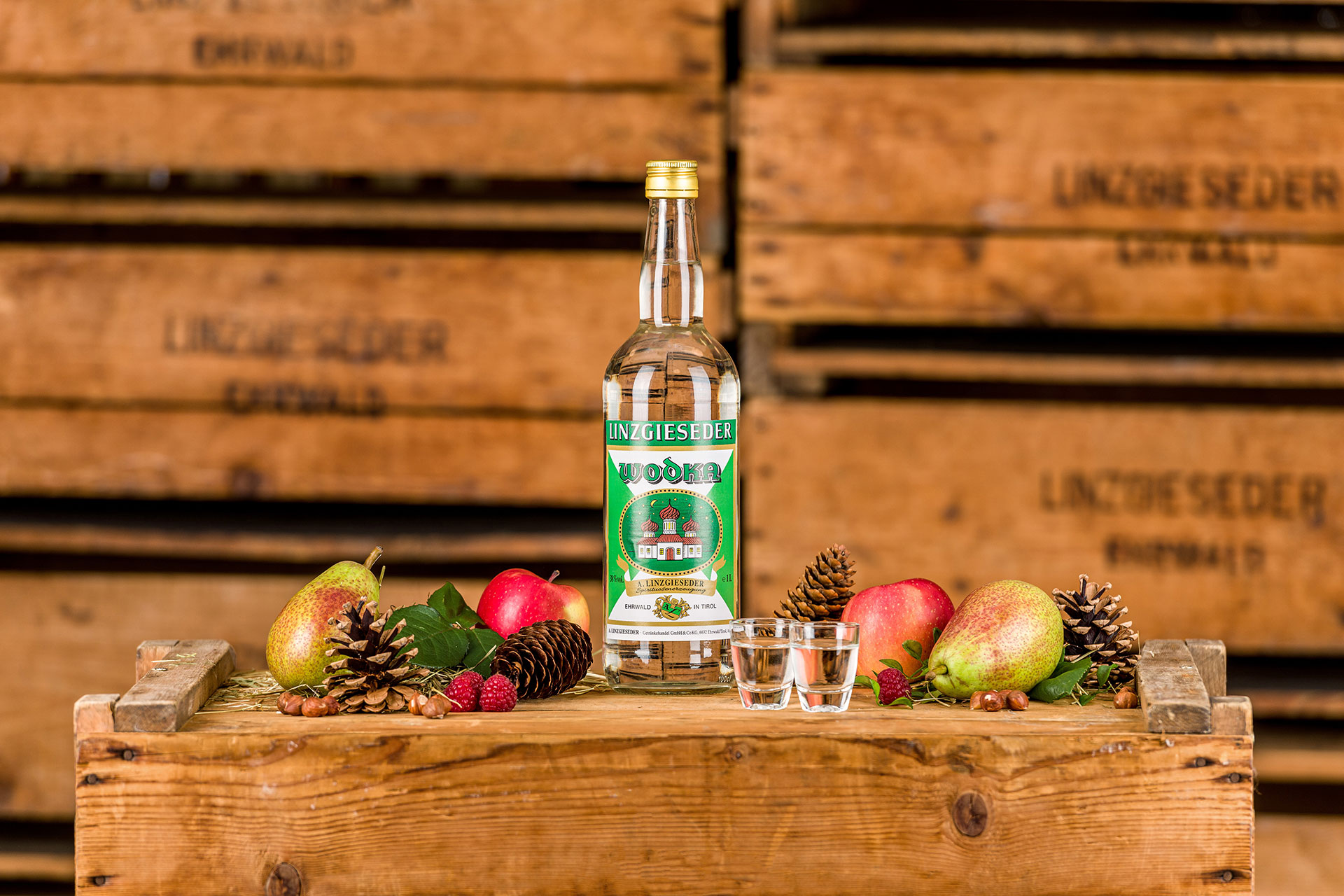 Spirituosenflasche Linzgieseder Wodka mit zwei gefüllten Schnapsgläsern mittig auf einer Holzkiste stehend. Links und rechts davon liegen Birnen, Äpfel, Zapfen und Nüsse. Der Hintergrund zeigt gestapelte Holzkisten mit Linzgieseder-Aufschrift.