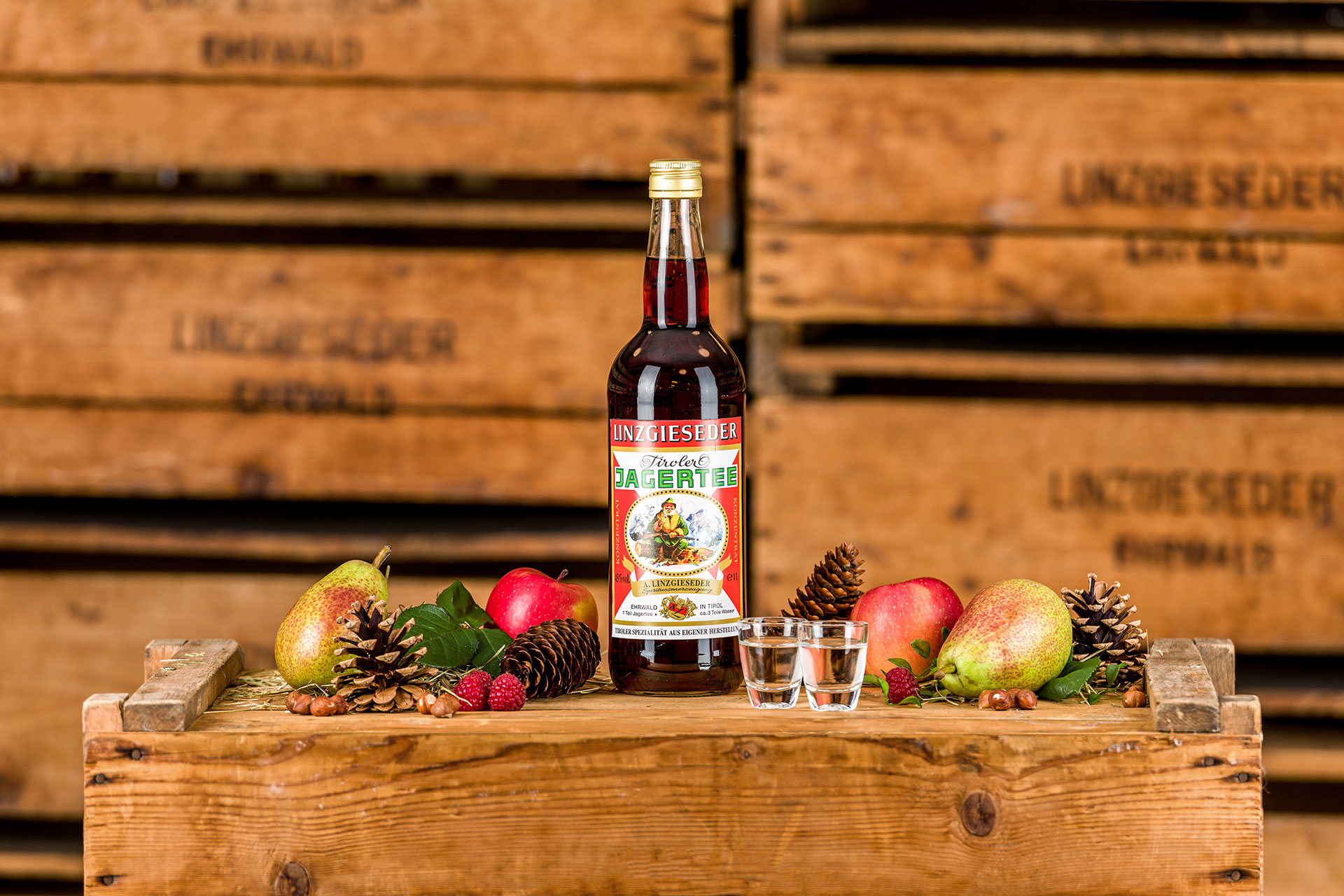 Spirituosenflasche Linzgieseder Jägertee 45% mit zwei gefüllten Schnapsgläsern mittig auf einer Holzkiste stehend. Links und rechts davon liegen Birnen, Äpfel, Zapfen und Nüsse. Der Hintergrund zeigt gestapelte Holzkisten mit Linzgieseder-Aufschrift.