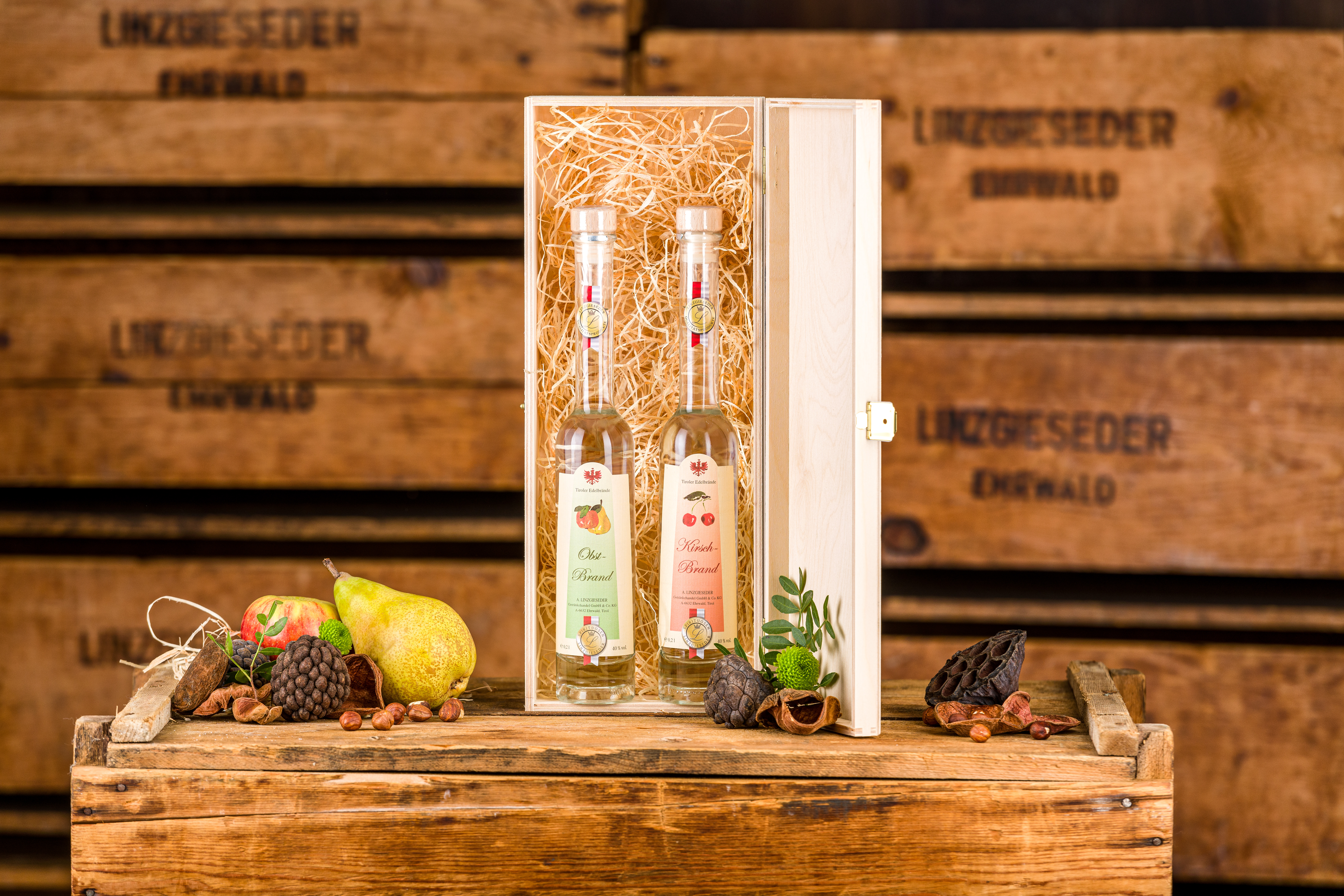 Zwei langhalsige Linzgieseder Edelbrandflaschen stehen auf einer Holzkiste. Dahinter befindet sich eine Geschenkverpackung in Form einer hellen Holzkiste mit strohartigem Füllmaterial, deren Deckel nach rechts geöffnet ist und die beiden Flaschen enthält.