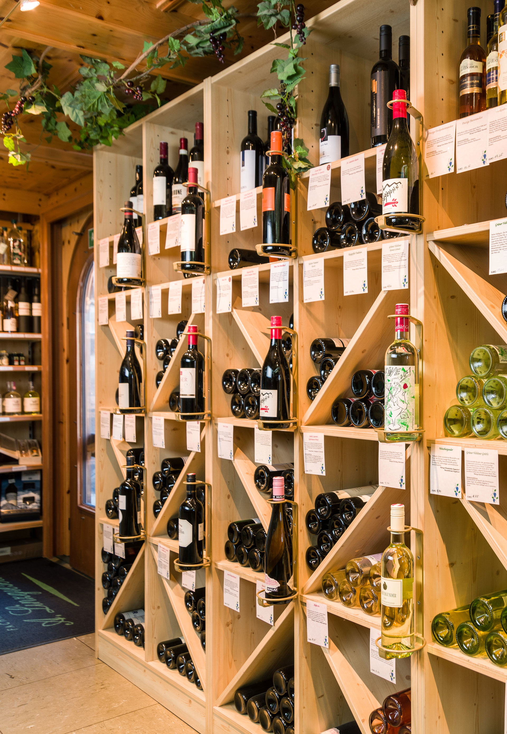 Das Bild zeigt die Weinregale aus der Schnaps- und Weinboutique in Ehrwald. Im Regal sind Rotwein- und Weißweinflaschen sichtbar, sowie eine Kunstweinpflanze, die von der Decke herunterhängt. Das Regal besteht aus hellem, freundlichem Holz und ist in mehr