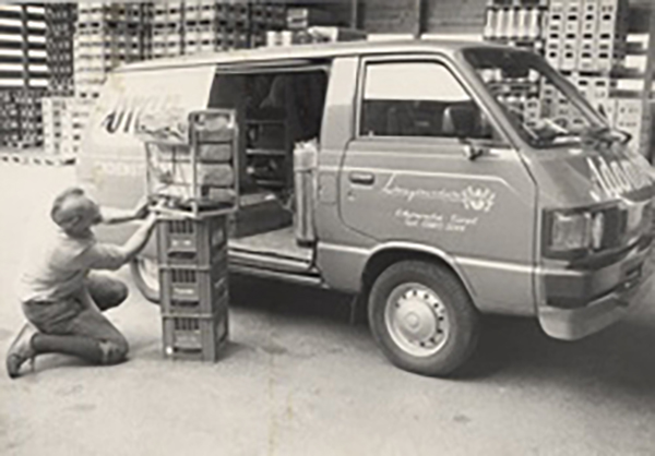 Ein schwarz-weißes Bild, auf dem im Mittelpunkt ein Opel Blitz mit offener Tür zu sehen ist. Im Hintergrund sind hohe Stapel von Getränkekisten und Paletten sichtbar. Vor der Seitentür des Opel Blitz kniet ein Mitarbeiter, vor ihm stehen Getränkekisten un