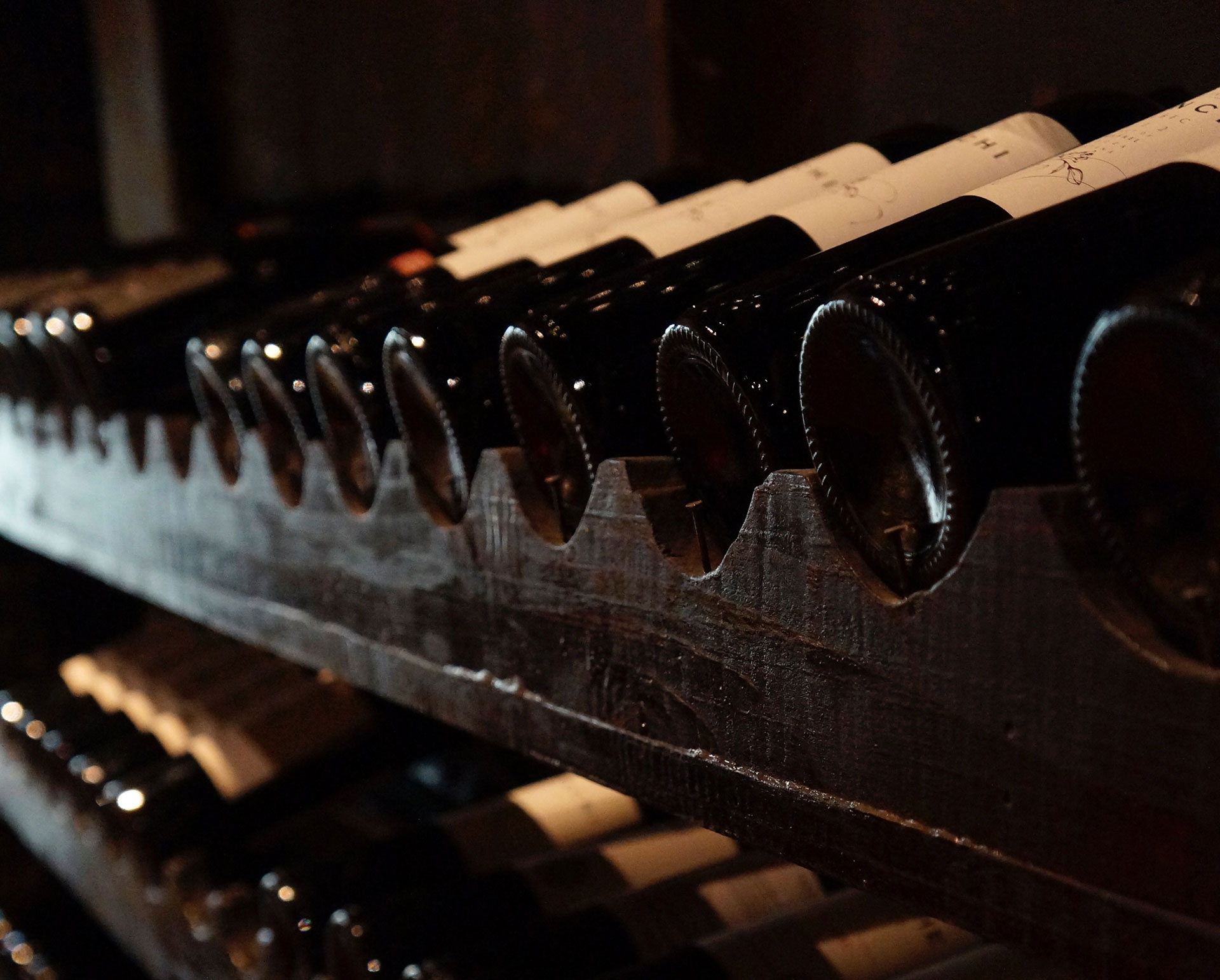 Nahaufnahme eines dunklen Weinregals, in dem Weinflaschen liegen. Nur die Böden der Flaschen und die hellen Etiketten sind sichtbar.