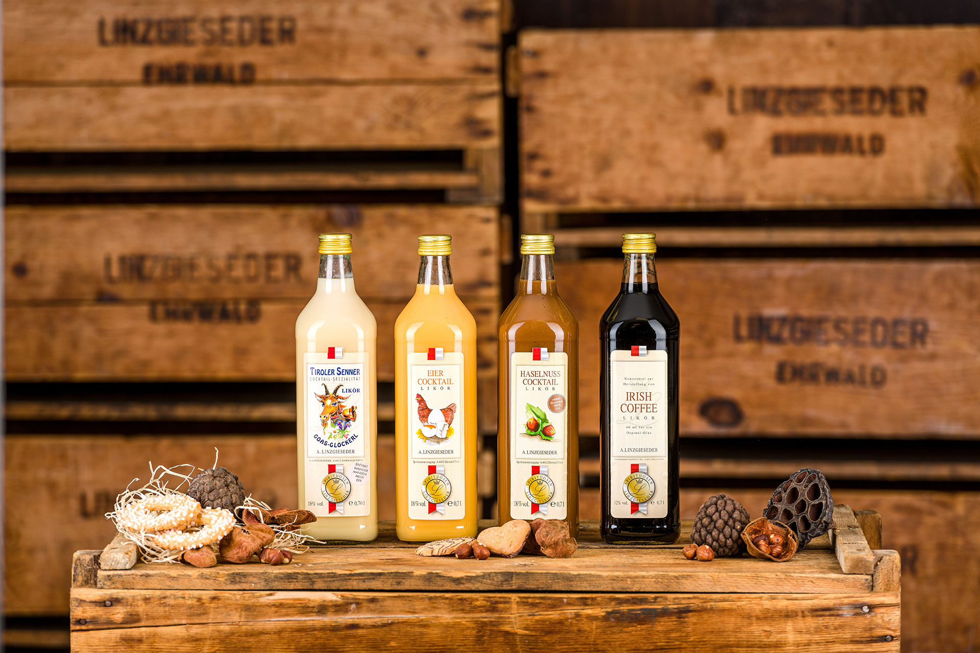 „Vier Flaschen auf einer Holzkiste stehend, von links nach rechts: Tiroler Senner in einer 0,75 l eckigen Flasche, Eier-Cocktail in einer 0,75 l eckigen Flasche, Haselnuss-Cocktail in einer 0,75 l eckigen Flasche und Irish Coffee Likör in einer 0,75 l eck