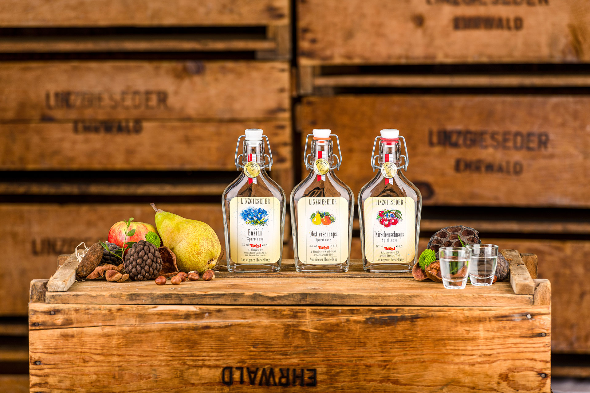 Drei Flaschen auf einer Holzkiste stehend, von links nach rechts: Enzian Spirituose in einer 0,35 l Bügelflasche, Obstlerschnaps in einer 0,35 l Bügelflasche und Kirschenschnaps in einer 0,35 l Bügelflasche. Links und rechts davon liegen Nüsse und Zapfen,