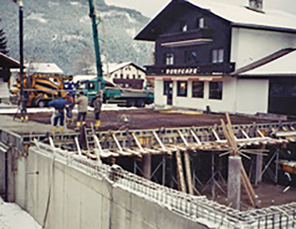 Auf dem Bild ist eine Großbaustelle zu sehen, bei der die Kellerdecke bereits halb fertiggestellt ist. Der Rohbau ist in vollem Gang. Im Hintergrund ist der alte Gasthof/Café sichtbar