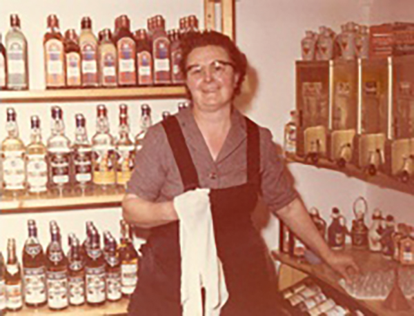 Eine lächelnde Frau steht vor einem Regal mit Linzgieseder Spirituosen, gekleidet im Stil des Jahres 1981. Mit der linken Hand greift sie nach einem Stamperl, während sie in der rechten ein Geschirrtuch hält