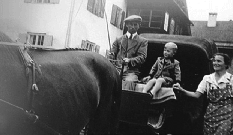 Eine schwarz-weiße Fotografie von Arnold Linzgieseder mit seinem Vater Alfred auf einem Pferdetaxi. Nur der Rücken des Pferdes ist erkennbar. Alfred Senior hält die Peitsche und die Zügel in der Hand. Die Mutter (Agnes) von Arnold geht rechts neben dem Pf