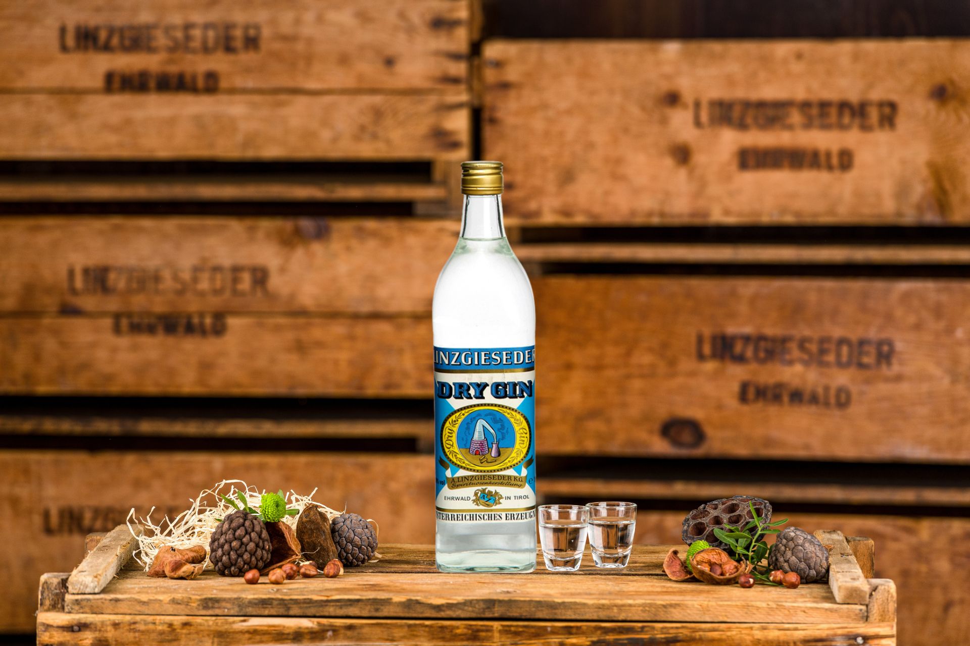 Spirituosenflasche Linzgieseder Gin mit zwei gefüllten Schnapsgläsern mittig auf einer Holzkiste stehend. Links und rechts davon liegen Zapfen und Nüsse. Der Hintergrund zeigt gestapelte Holzkisten mit Linzgieseder-Aufschrift.