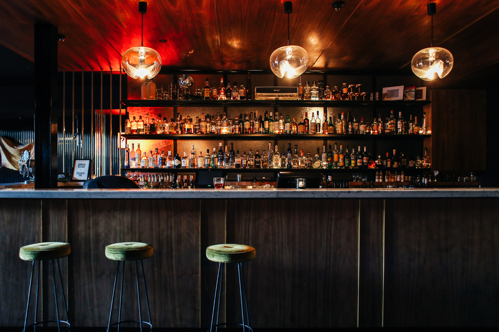 Blick auf eine stilvolle Bar mit drei Barhockern davor. Die Atmosphäre ist eher dunkel, und die Bar ist mit einer Vielzahl von Spirituosen bestückt. Drei Deckenlampen hängen herab und tauchen den Raum in ein sanftes Licht.