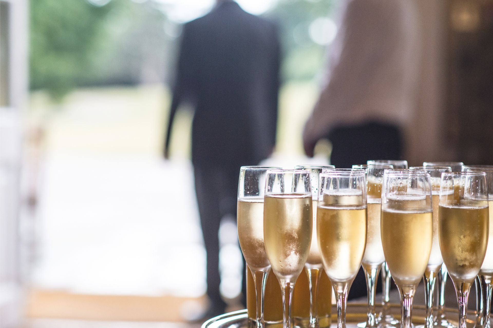 Ein Tablett mit gefüllten, goldgelben und leicht angelaufenen Sektgläsern im Vordergrund. Im Hintergrund sind verschwommen zwei Personen zu sehen, von denen eine einen Anzug trägt.