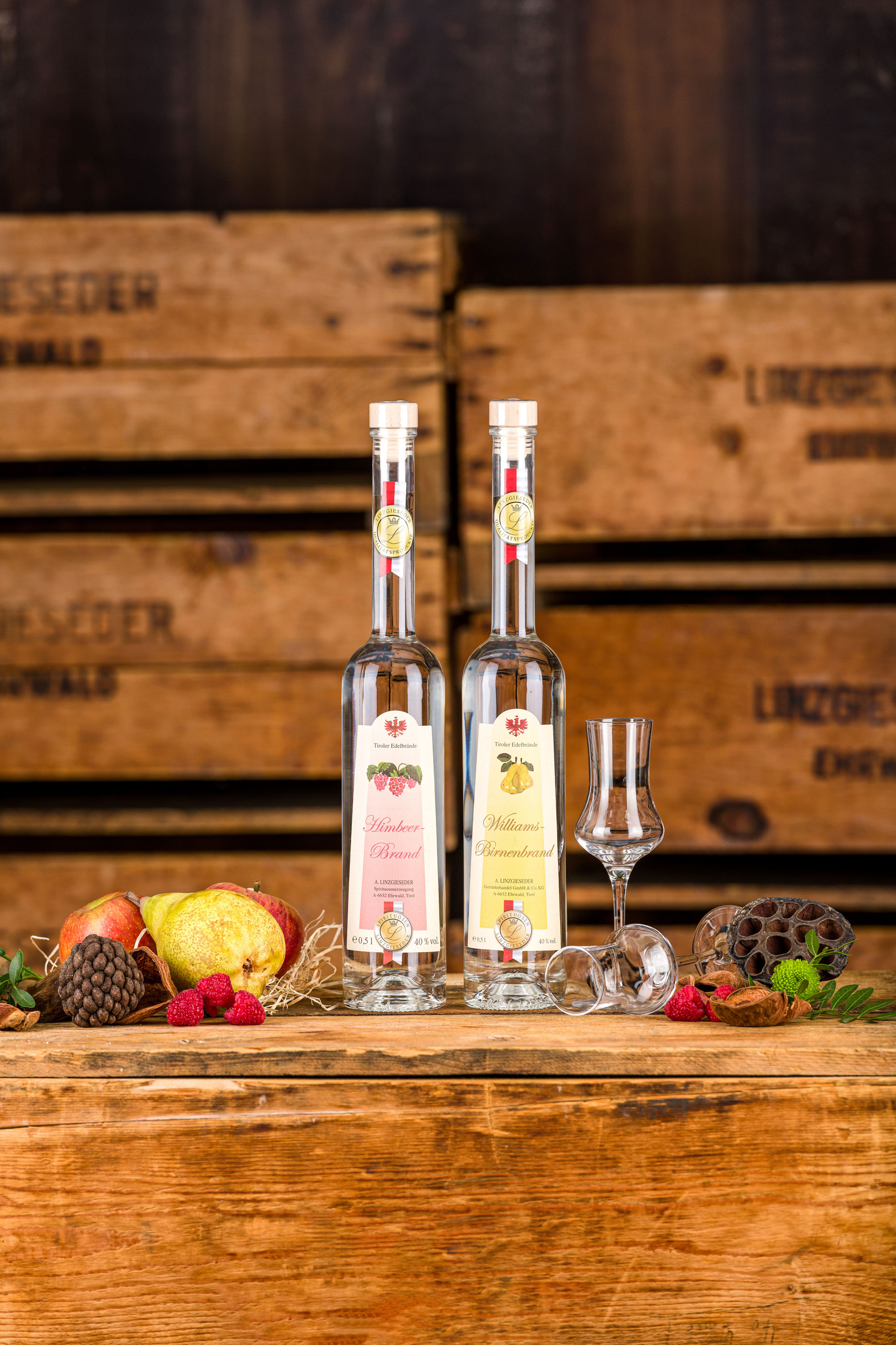 Zwei langhalsige Flaschen Linzgieseder Edelbrände mit zwei Selektionsgläsern, die mittig auf einer Holzkiste stehen. Ein Glas steht rechts neben den Flaschen, das andere liegt davor. Links und rechts davon liegen Birnen, Äpfel,Zapfen und Nüsse. Der Hinter