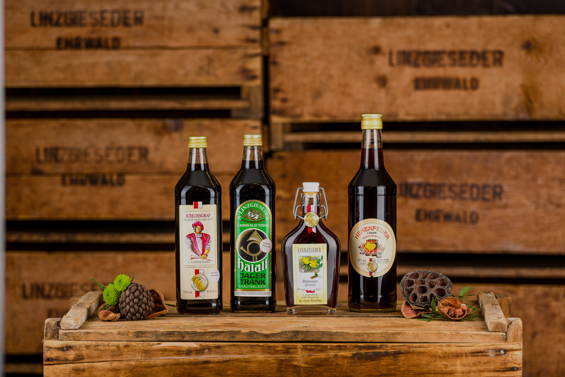 Vier Kräuterlikörflaschen auf einer Holzkiste stehend, von links nach rechts: Schlossgraf Likör in einer eckigen 0,75 l Flasche, Halali-Jagertrank in einer 0,75 l eckigen Flasche, Blutwurz in einer 0,5 l Bügelflasche und Hexenfeuer in einer 1 l runden Fla