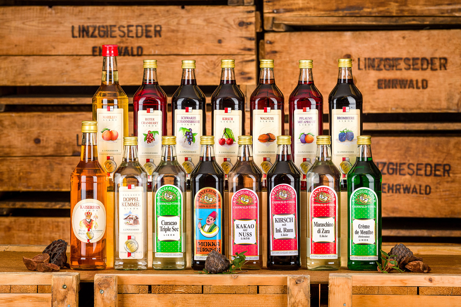 Bild mit 15 Likörflaschen, auf einer Holzkiste stehend, aufgeteilt in zwei Reihen: unten 8 Flaschen, dahinter auf einer erhöhten Stufe 7 Flaschen. Der Hintergrund zeigt Holzkisten mit Linzgieseder-Schriftzug